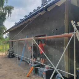 Dwuosobowa ekipa na rusztowaniu, montaż szarej izolacji termicznej na elewacji domu jednorodzinnego, widoczne paczki materiału Termo Organika, drabina przystawna z boku.
