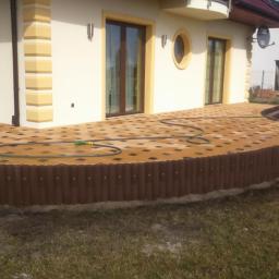 ROCKWOOD Zdzisław Gąsiorowski - Nowo ułożony, zakrzywiony taras z kostki brukowej w odcieniach brązu i beżu, otoczony niskim, ciemnobrązowym płotkiem z palików, z widocznym wężem ogrodowym na powierzchni, przylegający do jasnego...