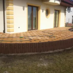 Nowo ułożony, zakrzywiony taras z kostki brukowej w odcieniach brązu i beżu, otoczony niskim, ciemnobrązowym płotkiem z palików, z widocznym wężem ogrodowym na powierzchni, przylegający do jasnego...