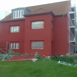 Czerwony dom w trakcie malowania elewacji, z oknami zabezpieczonymi folią i rusztowaniami. Na pierwszym planie elementy rusztowania i trawa.