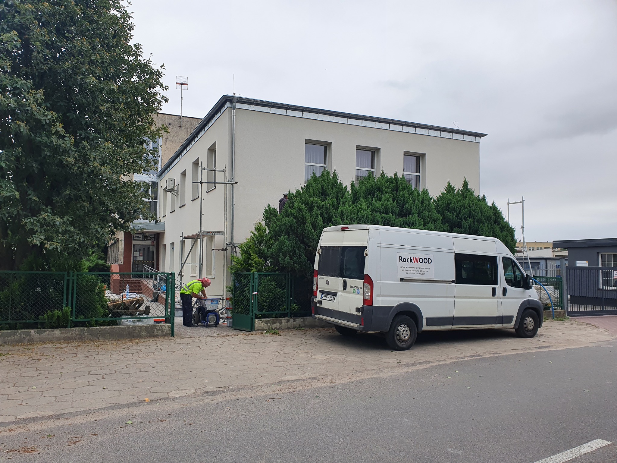 Pracownik w odblaskowej kamizelce przygotowuje maszynę do natrysku przy budynku z nową, jasną elewacją. W tle zaparkowany biały samochód dostawczy z logo Rockwool.