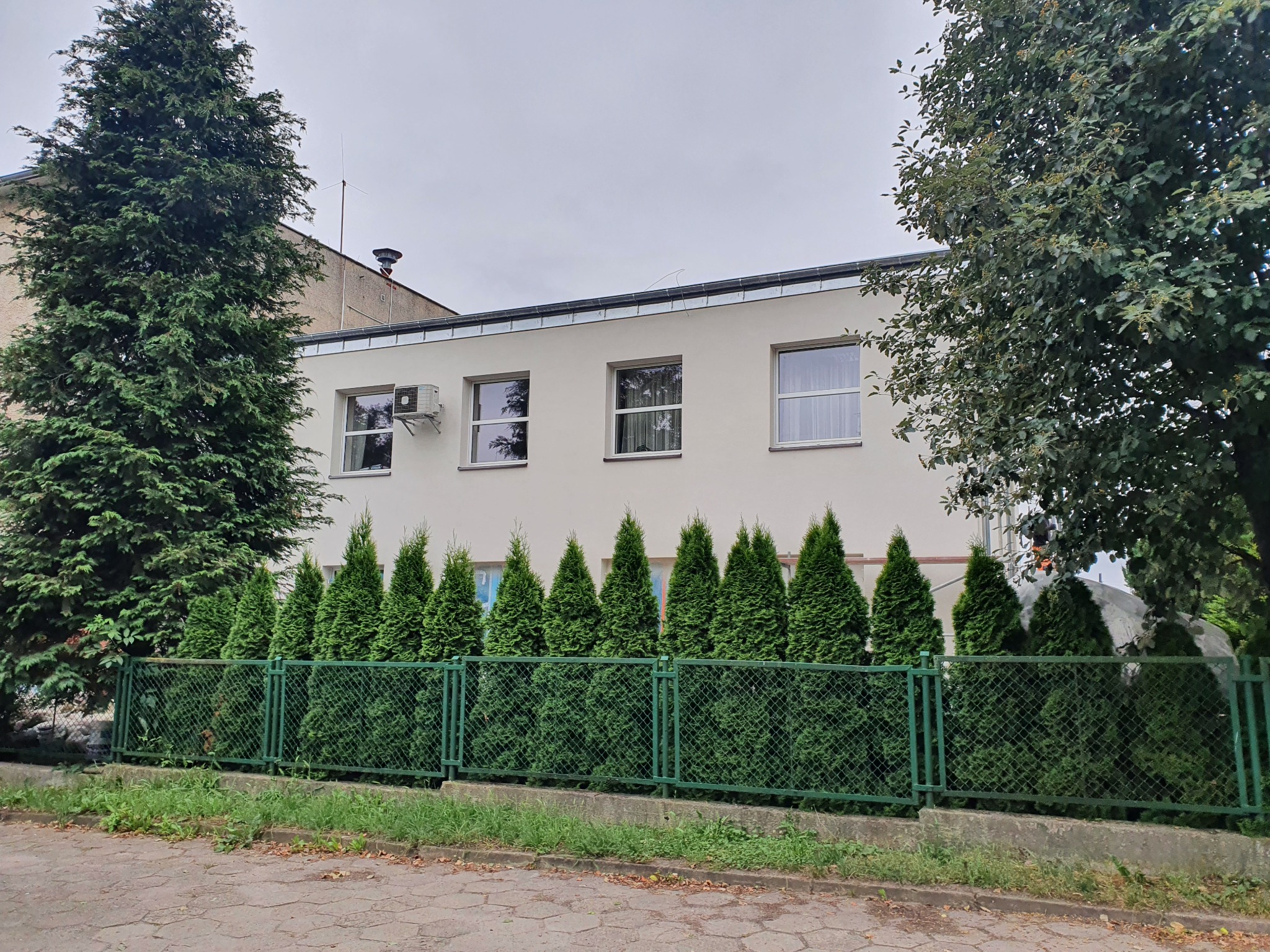 Jasna elewacja budynku z czterema oknami, klimatyzatorem i rzędem wysokich, zielonych tuj wzdłuż zielonego metalowego ogrodzenia.