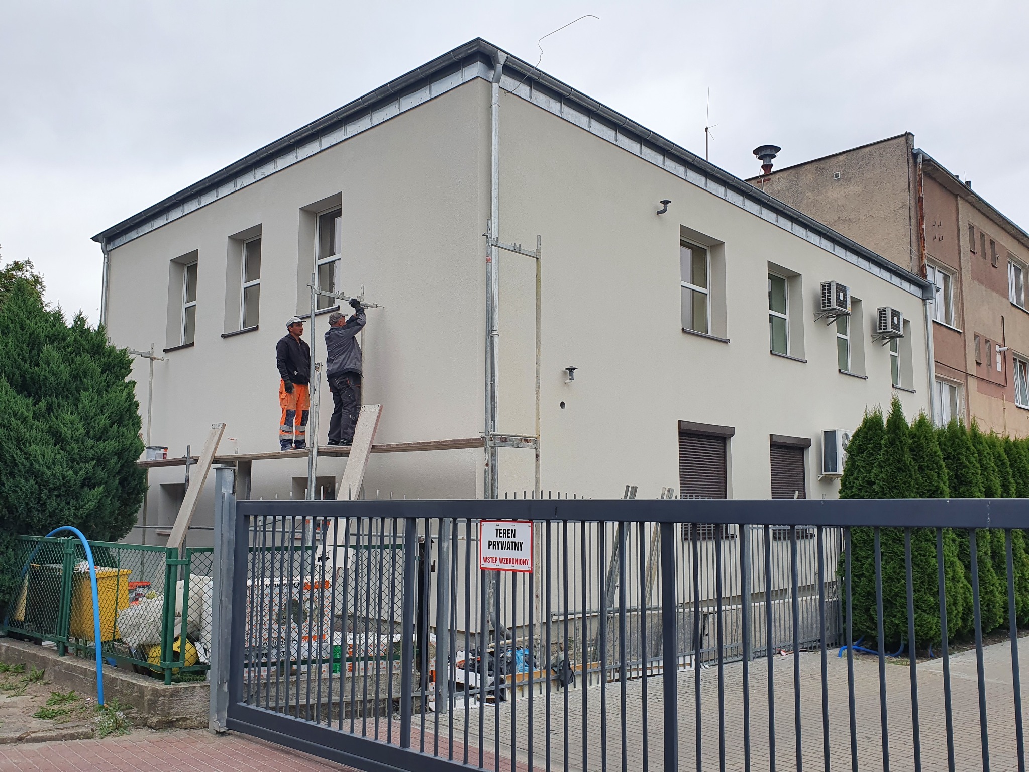 Dwóch pracowników na rusztowaniu przy dwukondygnacyjnym budynku w Przelewicach, jeden w kombinezonie roboczym, drugi w pomarańczowych spodniach, wykonuje prace wykończeniowe elewacji. W tle...