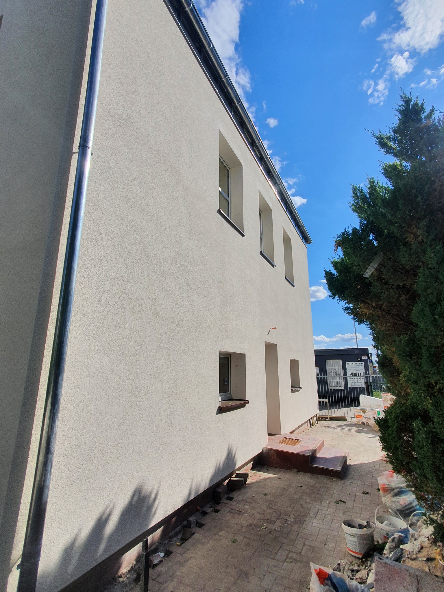 Jasna elewacja budynku z widocznymi oknami i rynnami, świeżo po nałożeniu tynku, z elementami wykończeniowymi w trakcie montażu, na tle błękitnego nieba z chmurami.