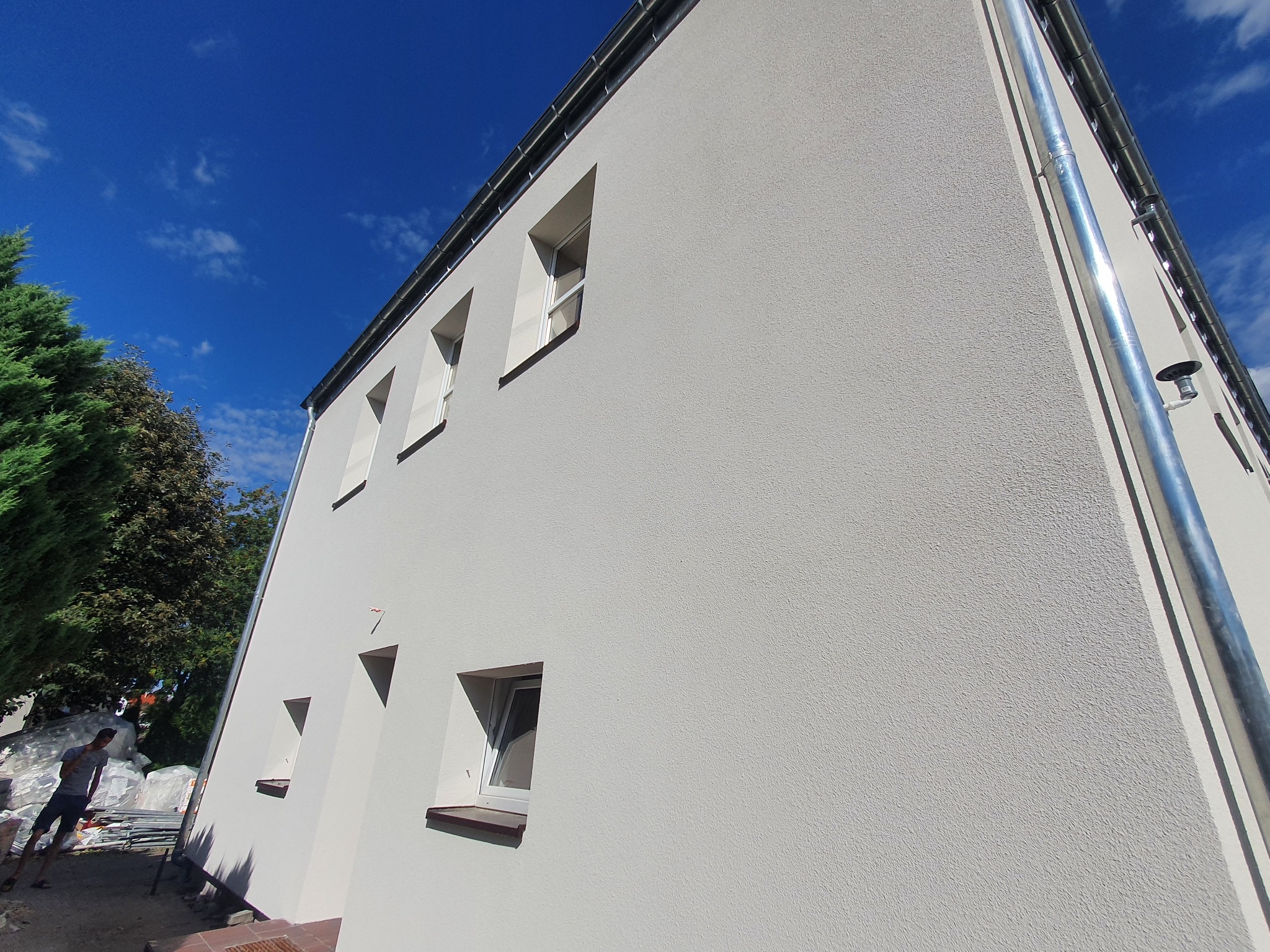 Świeżo otynkowana, jasna elewacja budynku z nietypowymi, trójkątnymi oknami, widok z dołu, błękitne niebo w tle, częściowo widoczny pracownik przy pracach wykończeniowych.
