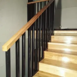 Jasne drewniane stopnie schod&oacute;w z czarną balustradą z pionowych szczebli i jasnym drewnianym poręczą, widoczne niedoskonałości na ścianie za balustradą.