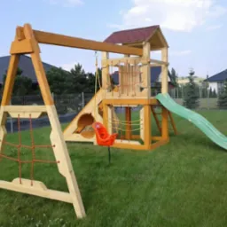 Drewniany plac zabaw z wieżą, zjeżdżalnią, huśtawką kubełkową i ścianką wspinaczkową z siatki, ustawiony na trawniku przed domem.