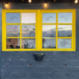 Okna w żółtej ramie, z naklejkami kwiatowymi na szybach, odbijające widok na morze i port, umieszczone na szarej drewnianej ścianie z wiszącą doniczką.