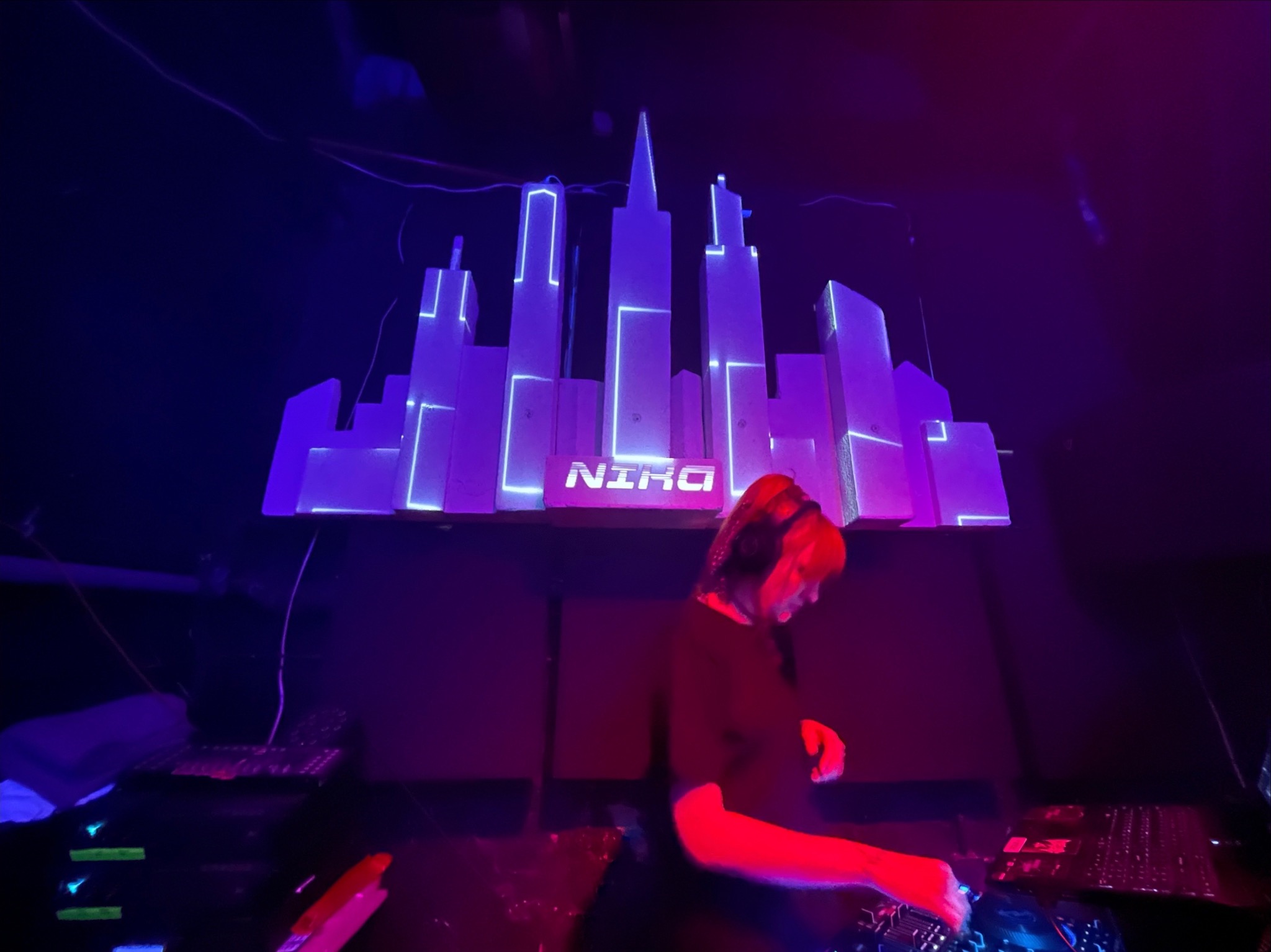 DJ-ka z rudymi włosami w słuchawkach miksuje muzykę przy konsolecie, w tle podświetlona makieta miasta z napisem 'NIRO'.