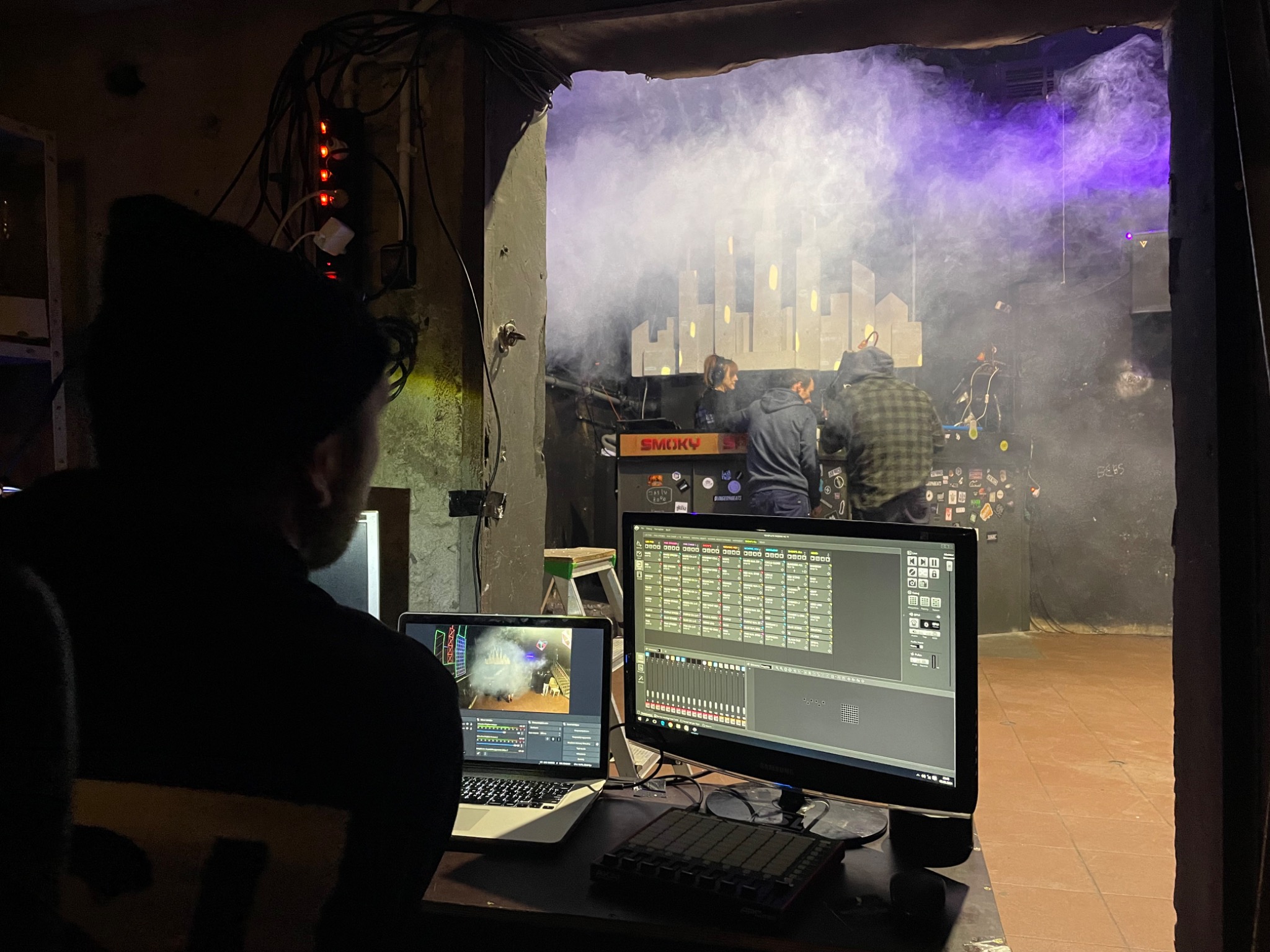 Osoba w czapce obserwuje scenę z dymem i podświetleniem, widoczną przez otwór w ścianie, z dwoma monitorami i kontrolerem MIDI przed sobą, ukazując oprogramowanie do edycji wideo i dźwięku.