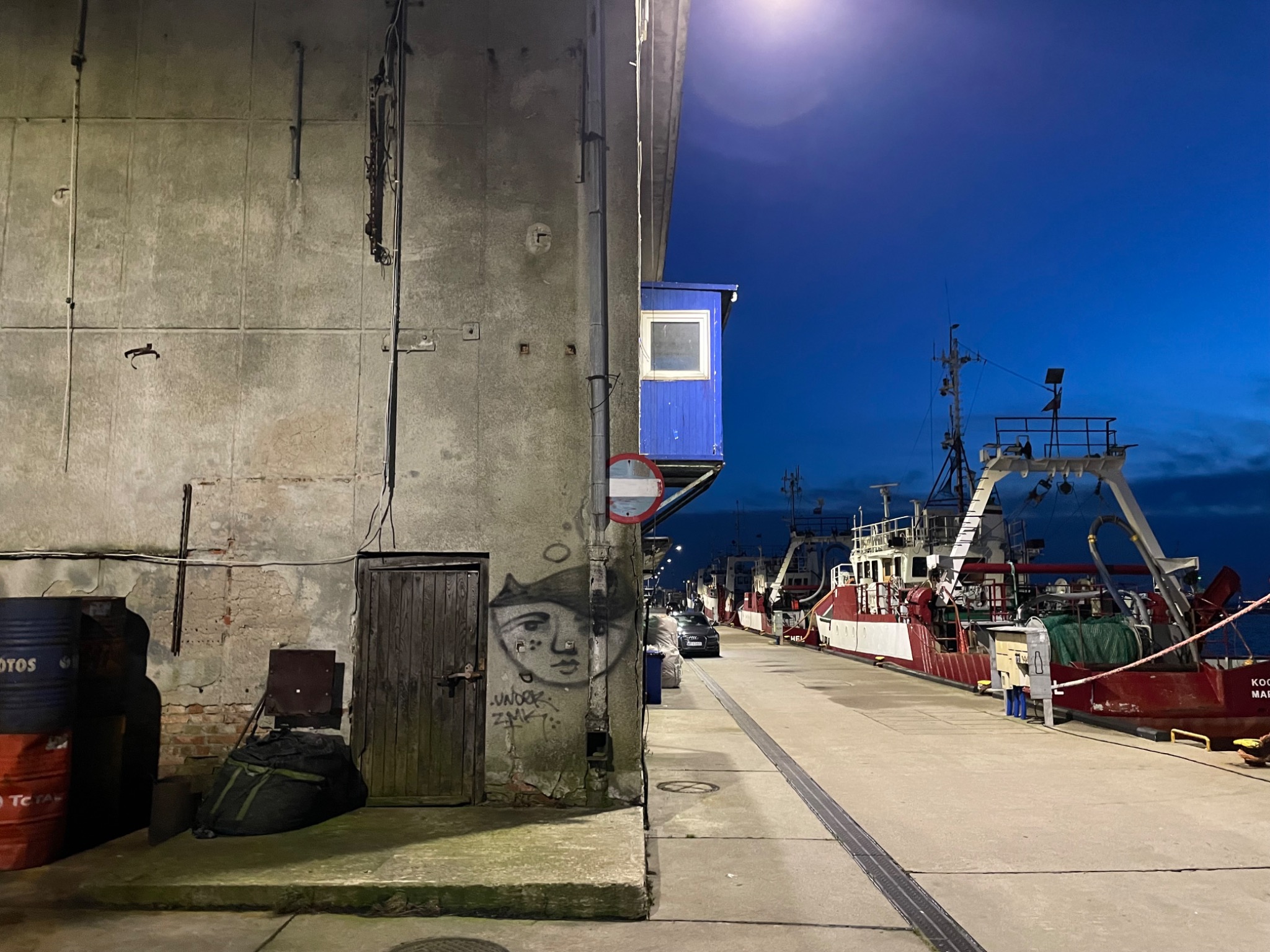 Nocny widok na port z betonową ścianą z graffiti i zaparkowanymi kutrami rybackimi w tle.