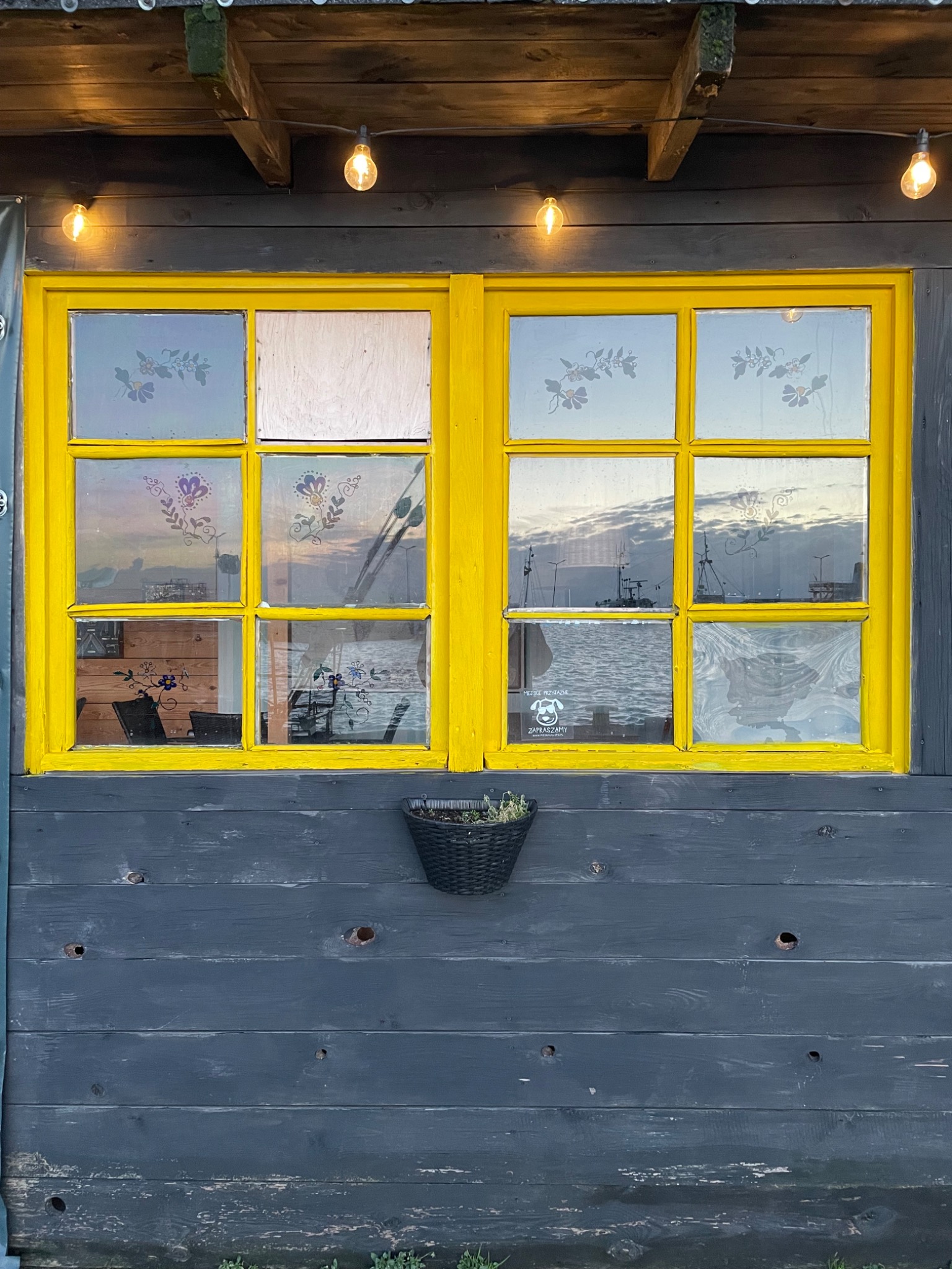Okna w żółtej ramie, z naklejkami kwiatowymi na szybach, odbijające widok na morze i port, umieszczone na szarej drewnianej ścianie z wiszącą doniczką.