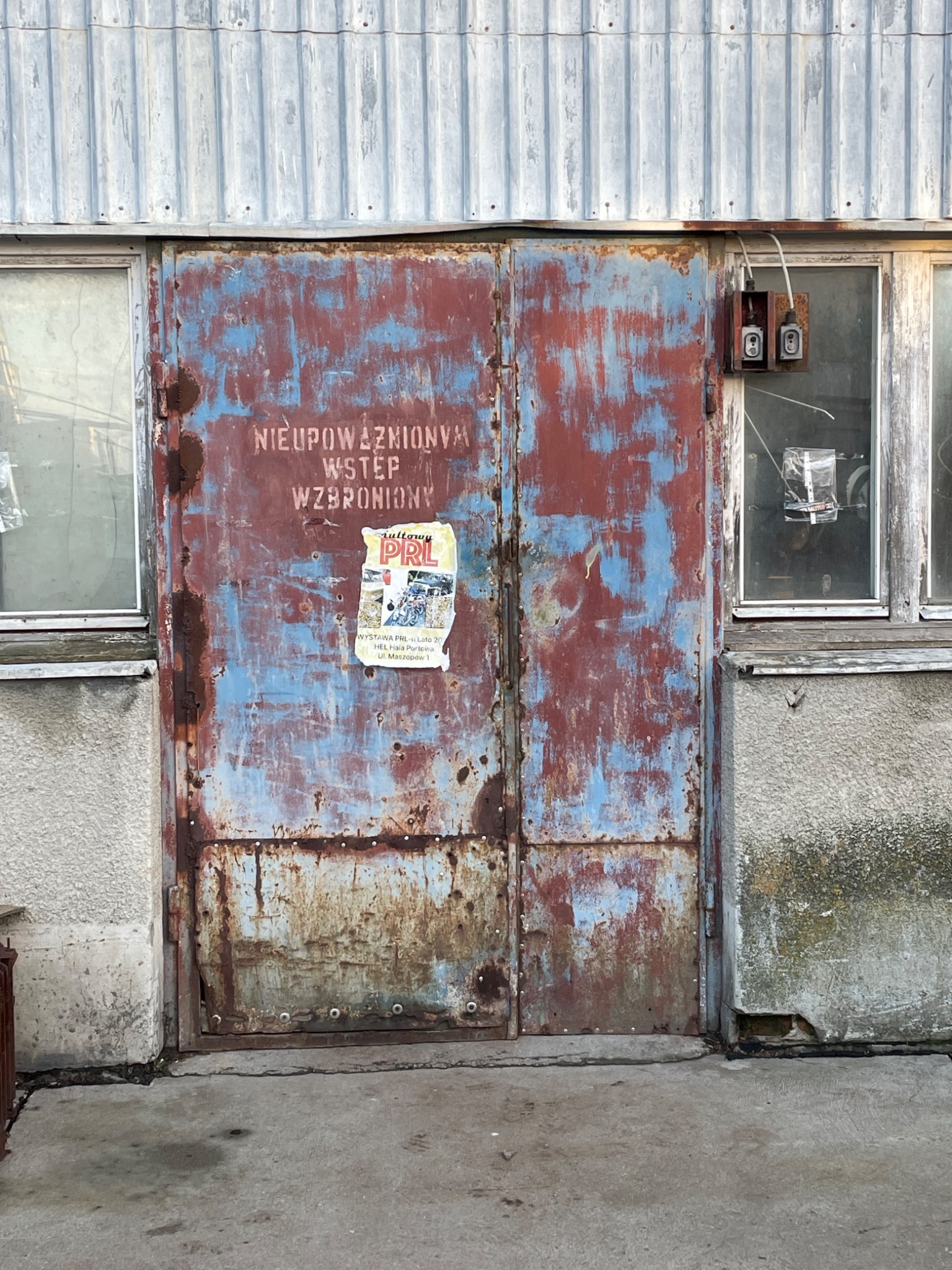 Zardzewiałe, dwuskrzydłowe metalowe drzwi z napisem 'NIEUPOWAŻNIONYM WSTĘP WZBRONIONY' i przyklejonym plakatem 'Kultowy PRL' wiszące na szarym, betonowym budynku z dwoma oknami i starymi gniazdkami...