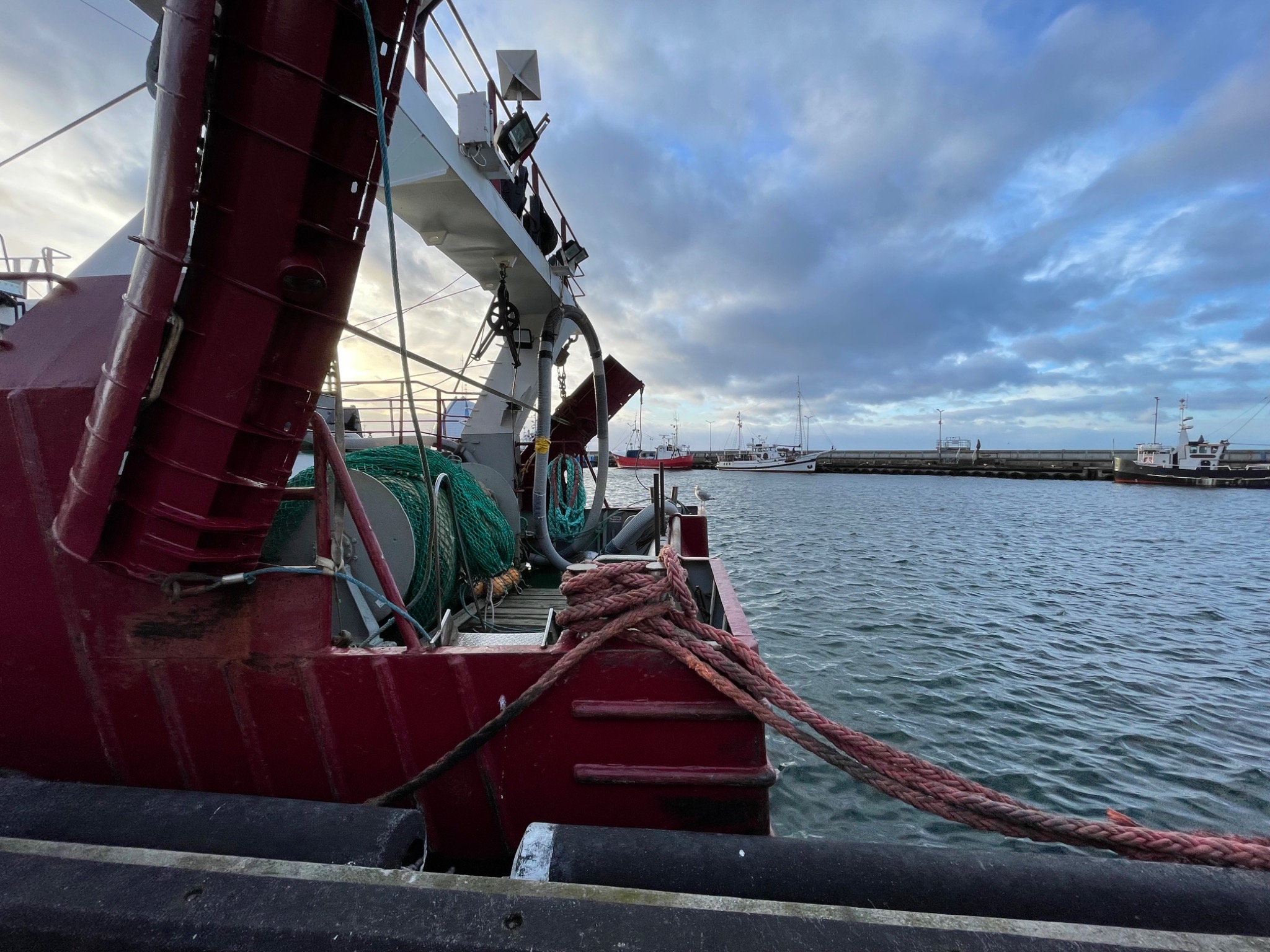 Część rufowa czerwonej łodzi rybackiej z grubymi linami na tle portu i pochmurnego nieba.