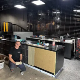 Pracownik w okularach klęczy przed ladą z częściowo położonymi czarnymi płytkami, widoczna warstwa kleju i płytki w rękach, czarne płytki na ścianie w tle.