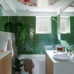 Jasna łazienka z zielonymi, błyszczącymi płytkami, okrągłym panelem z motywem kwiatowym na suficie, wiszącymi roślinami w doniczkach i drewnianymi szafkami pod umywalką.