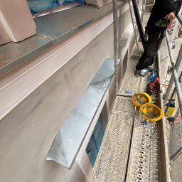 Oleksandr Kolisnyk - Montaż nowego metalowego parapetu zewnętrznego na elewacji budynku z rusztowania, widoczne narzędzia i kaski pracowników.