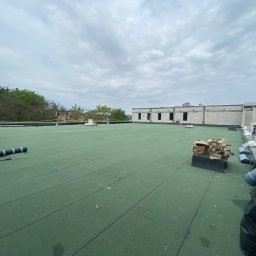 Oleksandr Kolisnyk - Zielony, płaski dach pokryty papą termozgrzewalną w trakcie prac dekarskich, widoczne rolki papy, narzędzia dekarskie i fragment budynku w surowym stanie na tle pochmurnego nieba.