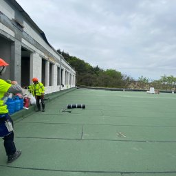 Oleksandr Kolisnyk - Prace dekarskie na płaskim dachu budynku w trakcie budowy: robotnicy w odblaskowych ubraniach i kaskach układają zieloną papę termozgrzewalną, widoczne butle gazowe i narzędzia dekarskie.