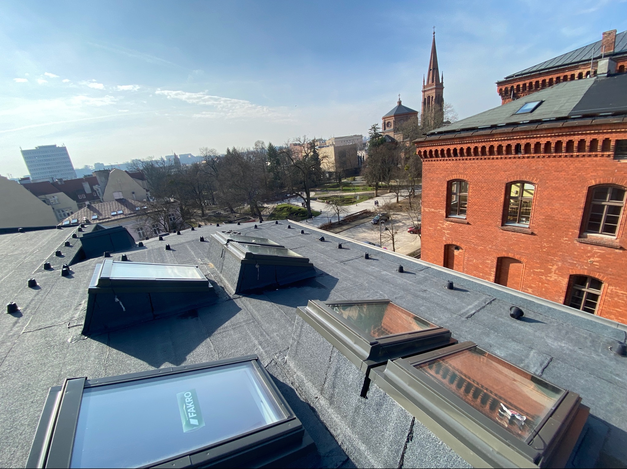 Widok z dachu pokrytego papą na miasto z kościołem i budynkiem z czerwonej cegły; widoczne okna dachowe Fakro z folią ochronną.