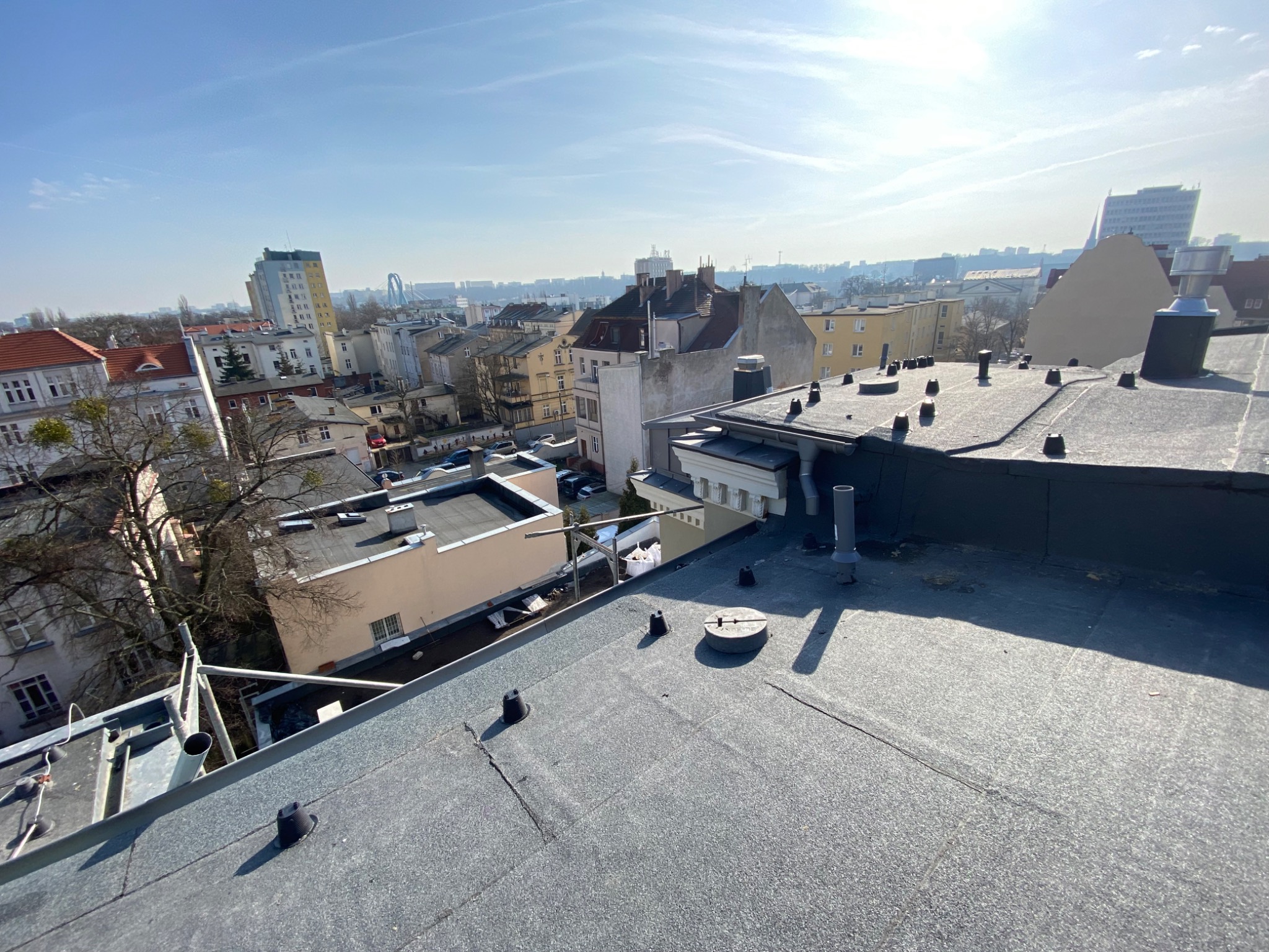 Widok z góry na płaski dach pokryty papą termozgrzewalną z widocznymi elementami wentylacyjnymi i kominami na tle zabudowy miejskiej w słoneczny dzień.