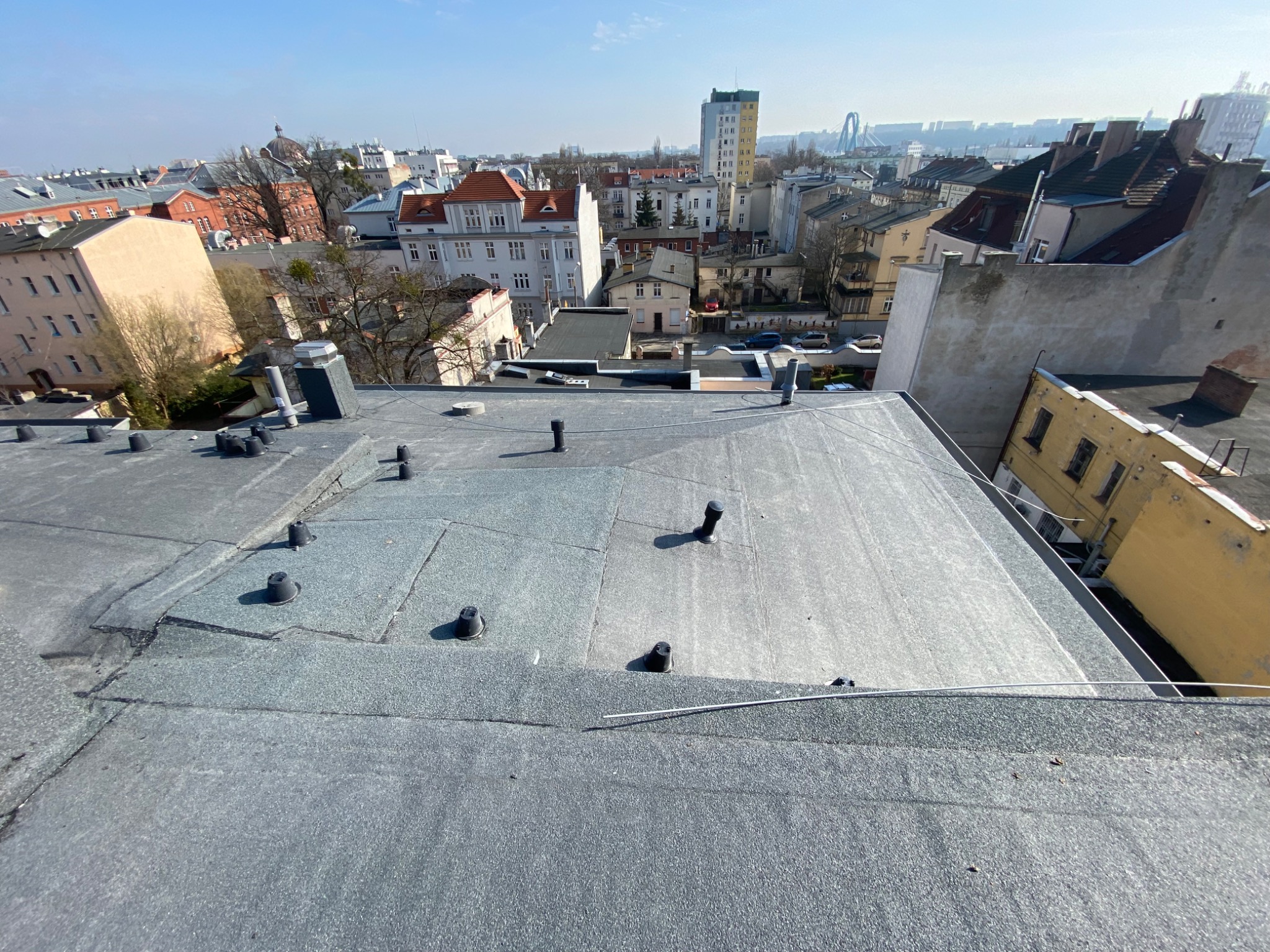 Płaski dach pokryty papą termozgrzewalną z widocznymi kominkami wentylacyjnymi na tle zabudowy miejskiej w słoneczny dzień.