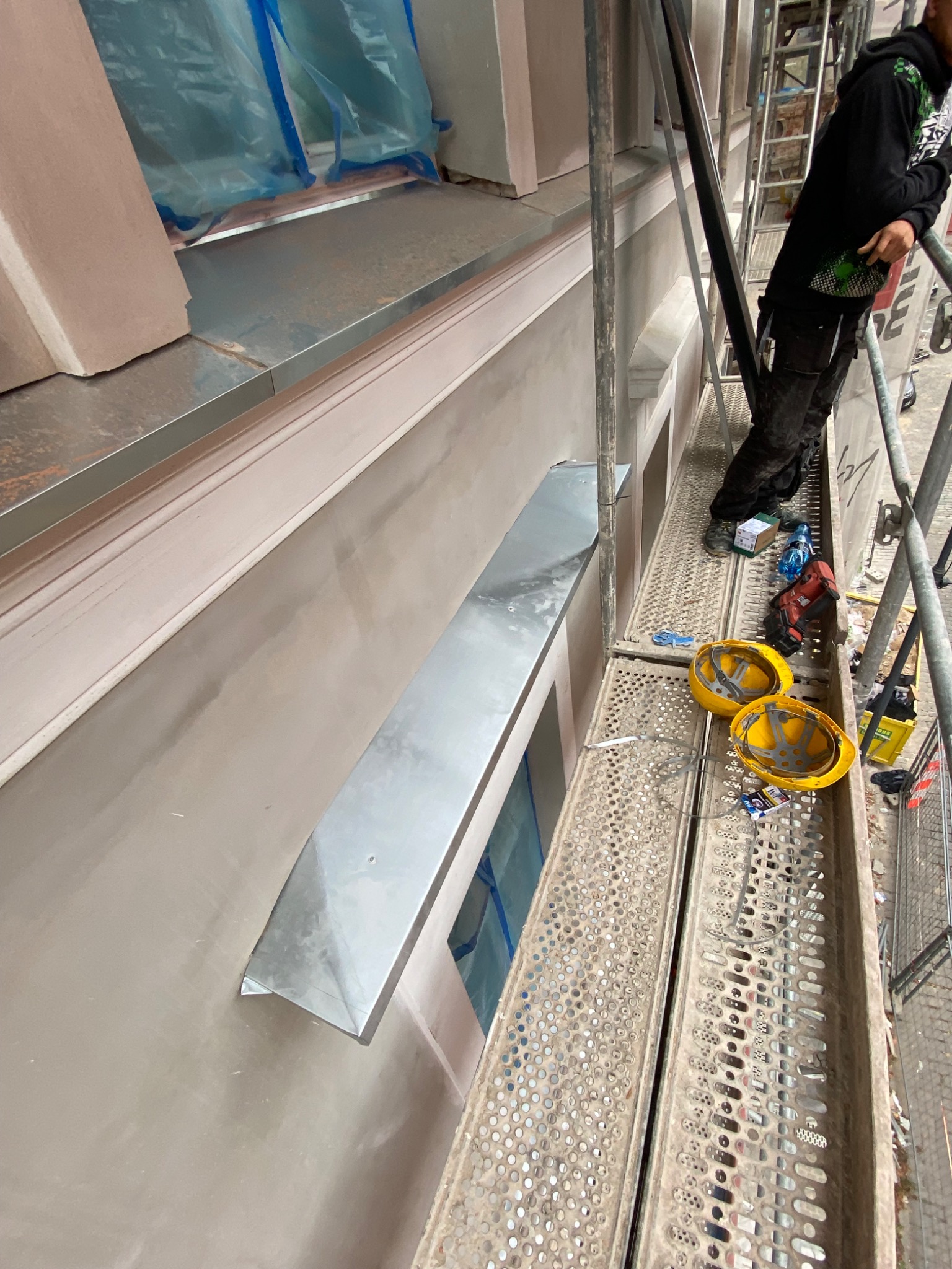 Montaż nowego metalowego parapetu zewnętrznego na elewacji budynku z rusztowania, widoczne narzędzia i kaski pracowników.