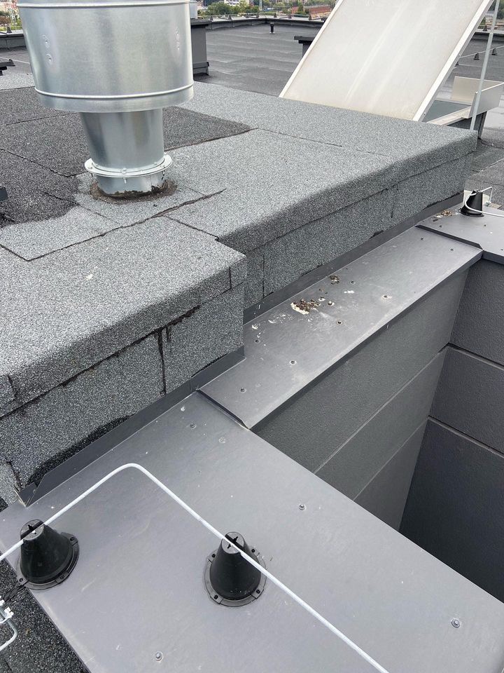 Płaski dach pokryty papą z kominem wentylacyjnym i zabezpieczeniami odgromowymi, widoczne detale połączeń i wykończeń.