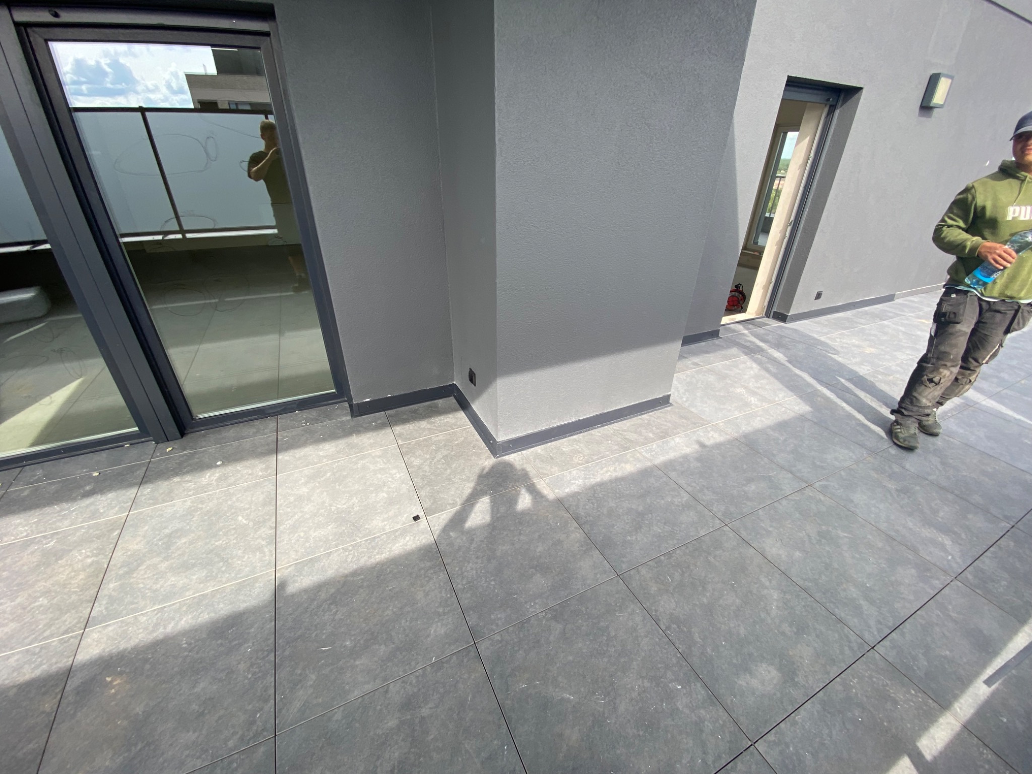 Szare płytki na balkonie nowoczesnego budynku, widoczny pracownik w ubraniu roboczym z butelką wody, szara elewacja budynku z oknami i drzwiami balkonowymi.