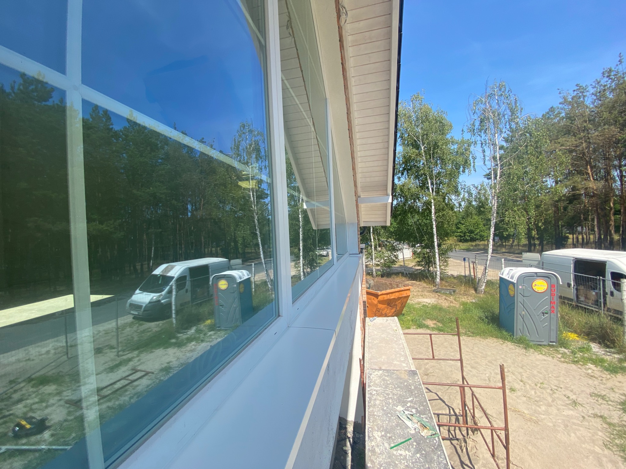 Widok z rusztowania na nowo zamontowane okna i fragment dachu z obróbkami blacharskimi, w tle las i samochody dostawcze.