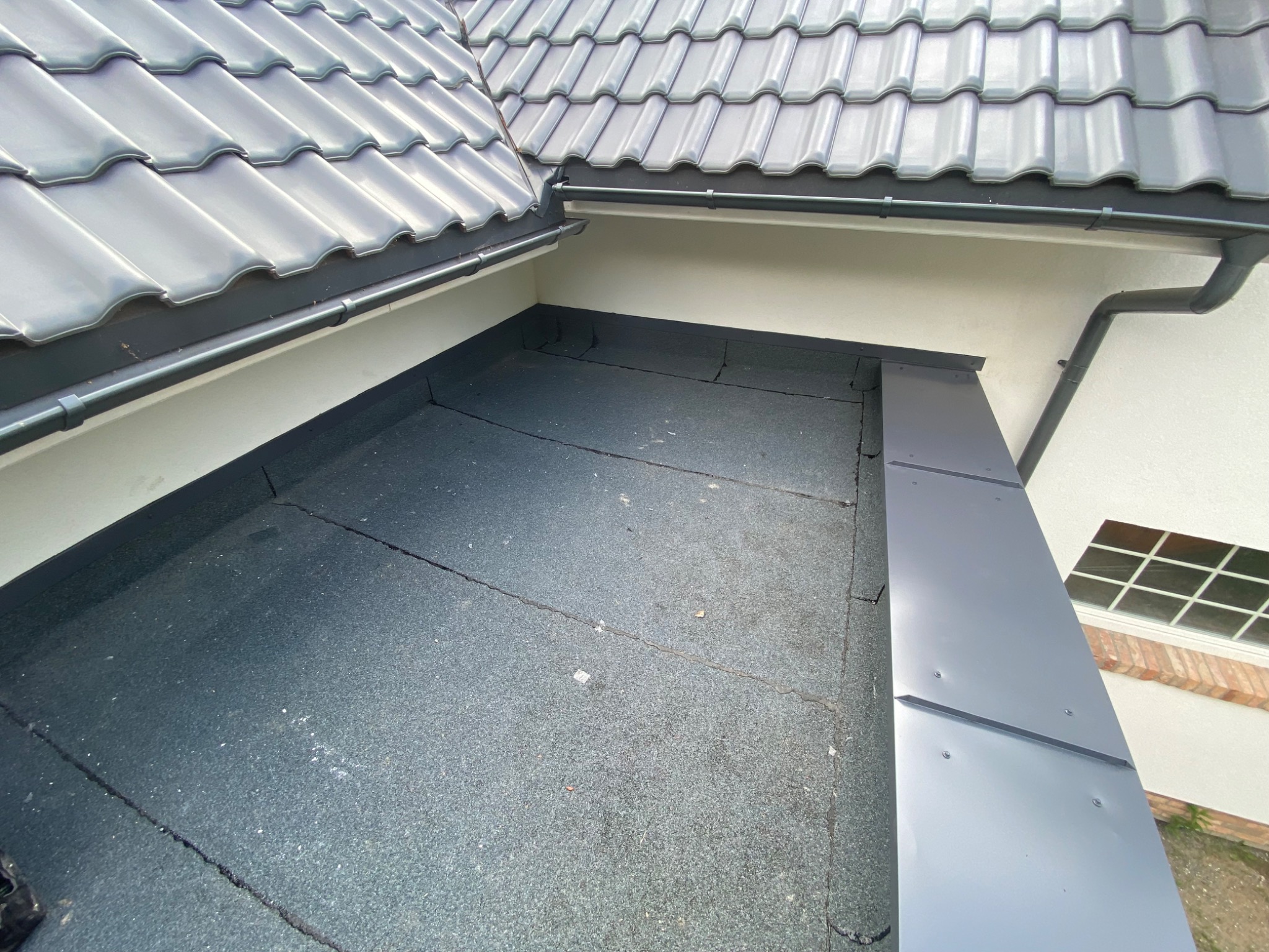 Widok z góry na fragment dachu z szarymi dachówkami i papą, z widocznymi rynnami i obróbkami blacharskimi na krawędziach.