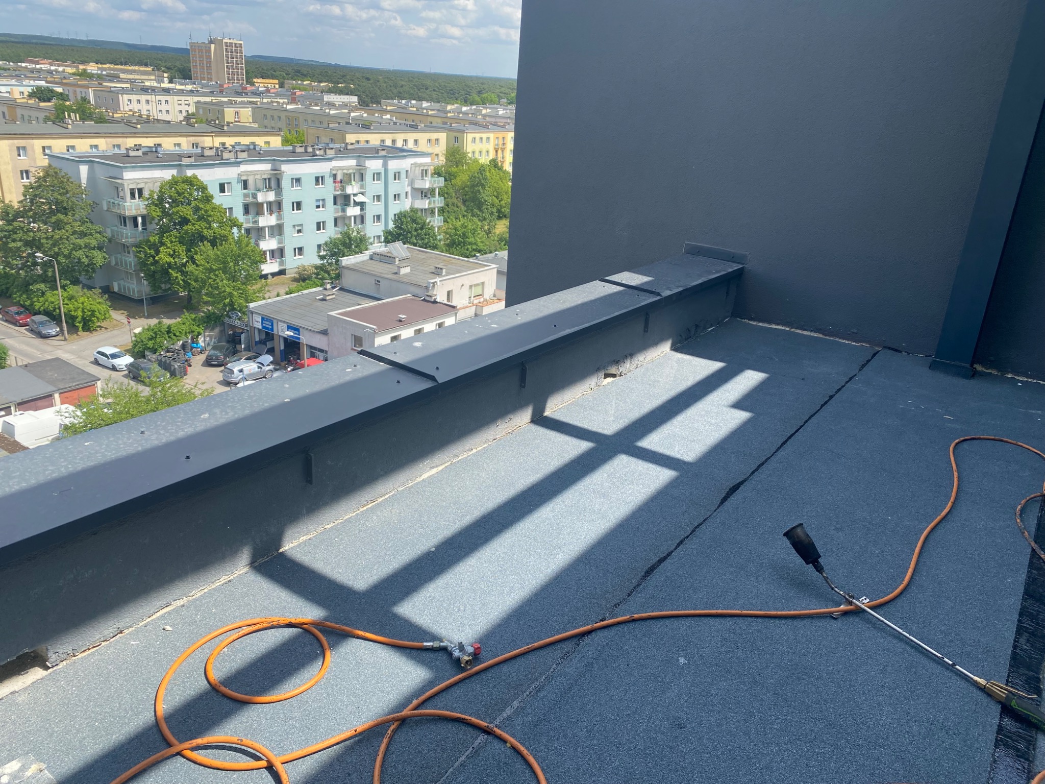 Prace na dachu płaskim z użyciem palnika dekarskiego, widoczne zgrzewane pasy papy i cień konstrukcji na tle zabudowy miejskiej.