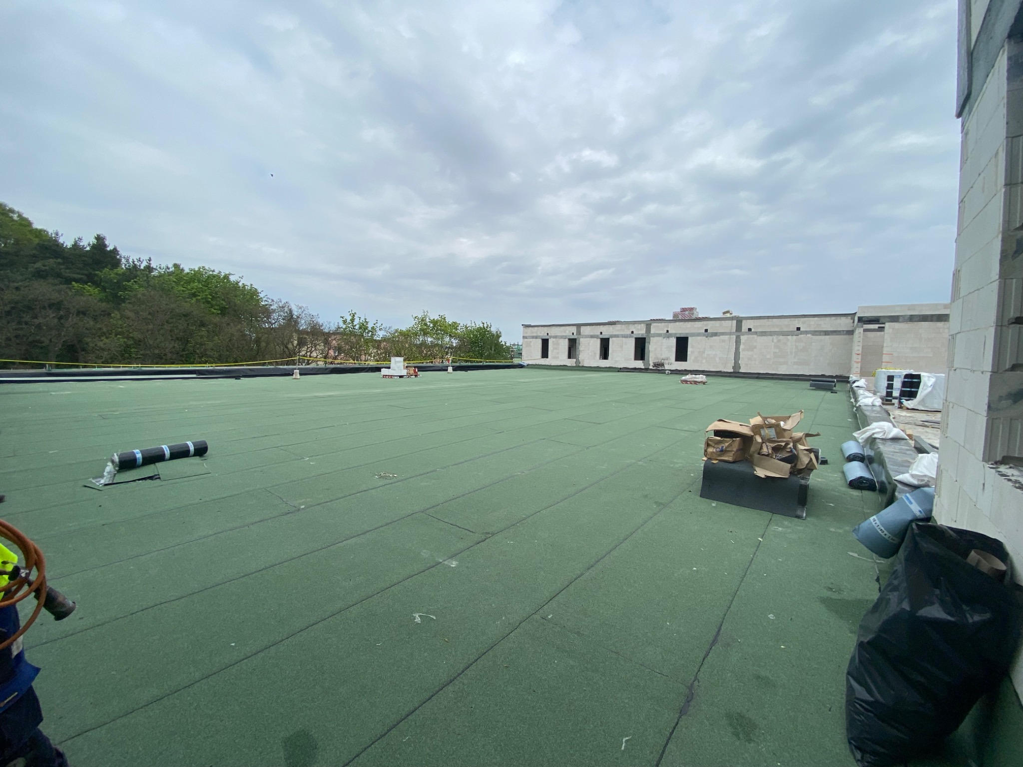 Zielony, płaski dach pokryty papą termozgrzewalną w trakcie prac dekarskich, widoczne rolki papy, narzędzia dekarskie i fragment budynku w surowym stanie na tle pochmurnego nieba.