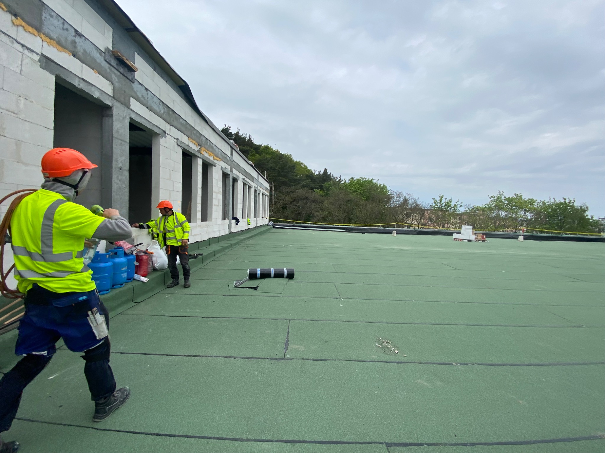 Prace dekarskie na płaskim dachu budynku w trakcie budowy: robotnicy w odblaskowych ubraniach i kaskach układają zieloną papę termozgrzewalną, widoczne butle gazowe i narzędzia dekarskie.