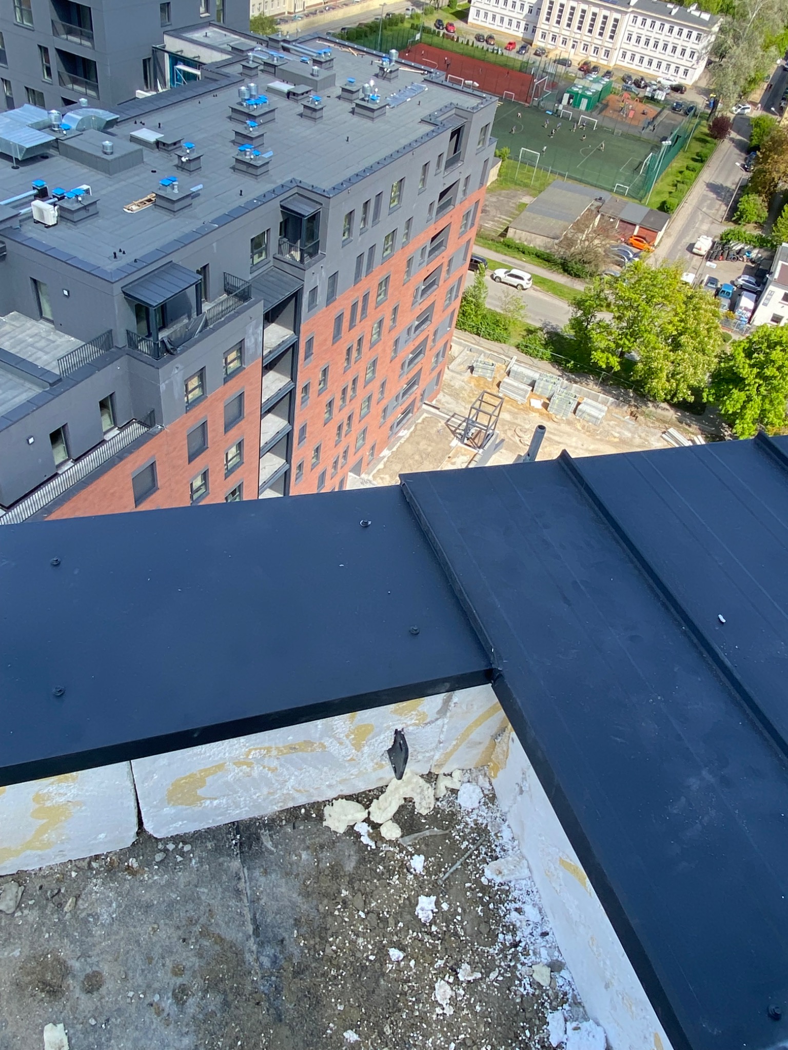 Widok z góry na fragment nowo położonego, czarnego arkusza blachy na dachu budynku wielorodzinnego, z widocznymi elementami mocującymi i fragmentem muru oraz panoramą miasta w tle.