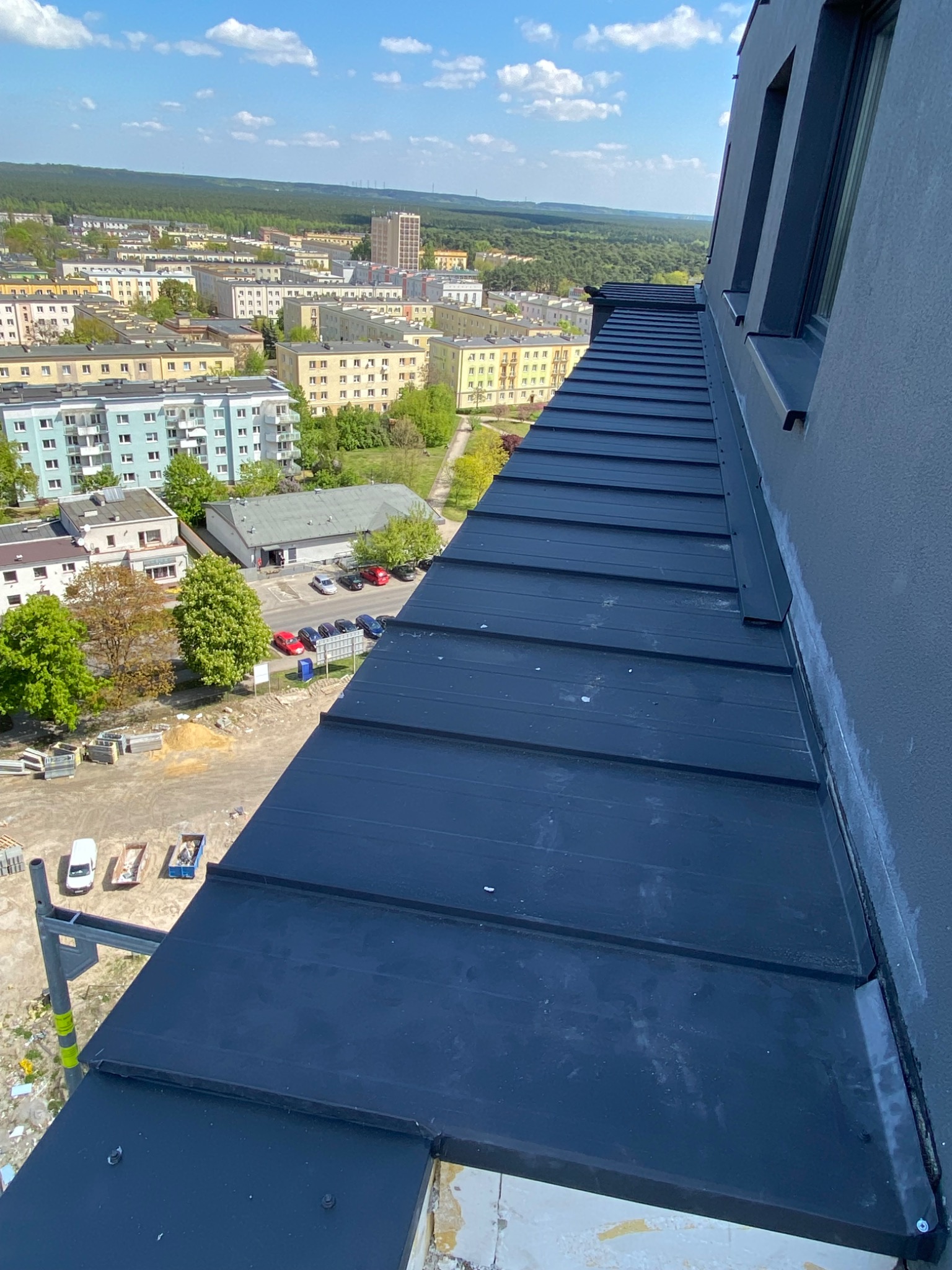 Widok z góry na nowo położony, czarny dach z blachy na budynku w Bydgoszczy, z widocznym fragmentem elewacji i panoramą miasta w tle.