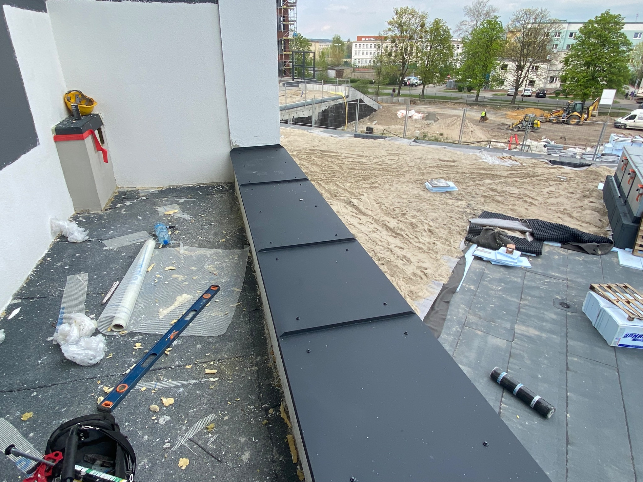 Montaż czarnych paneli na attyce dachu, widoczne narzędzia, poziomica, resztki materiałów izolacyjnych i budowa w tle.