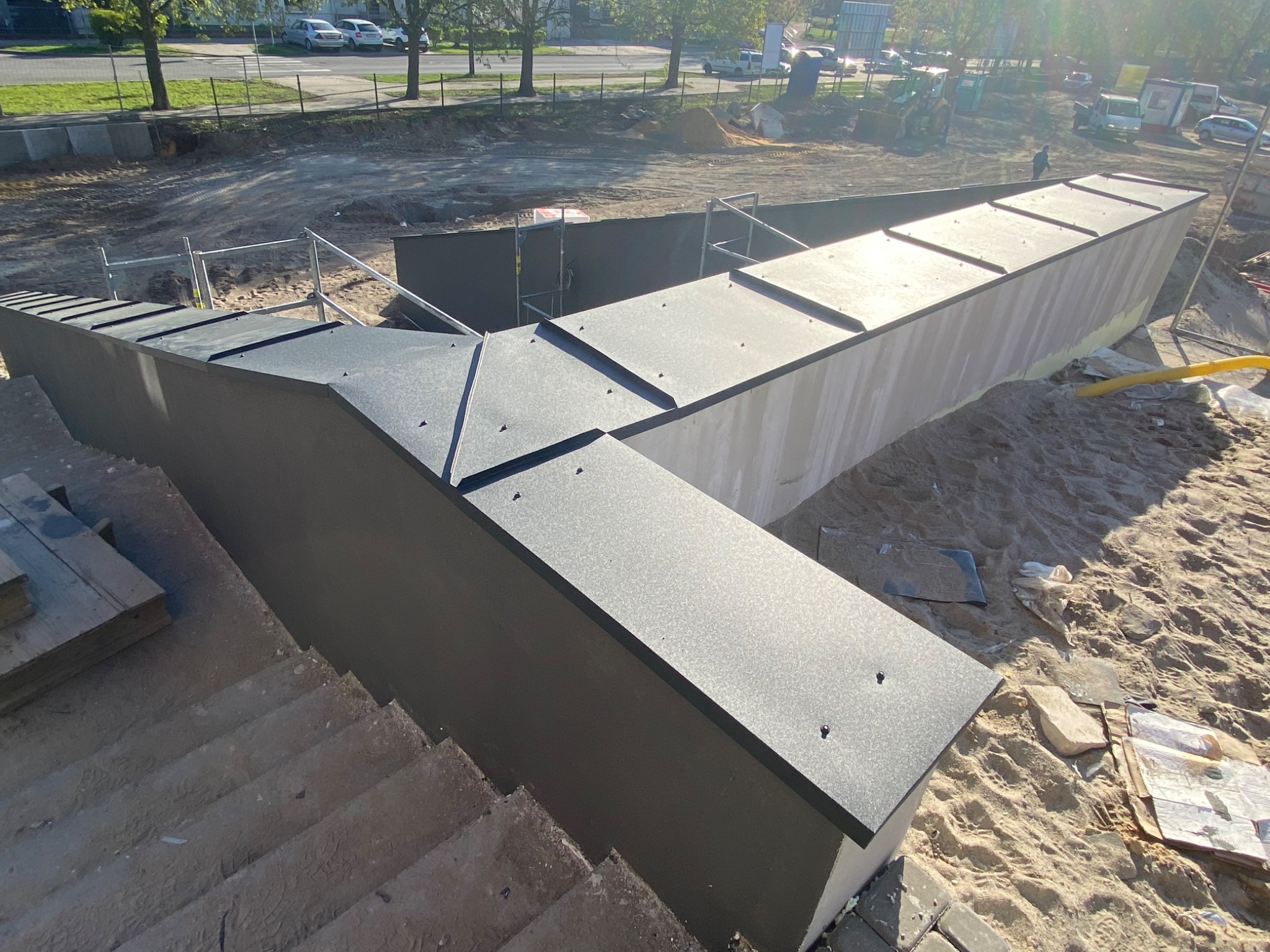 Nowoczesna konstrukcja z blachy trapezowej w kształcie litery V, prawdopodobnie element odwodnienia liniowego, na placu budowy z piaskiem i widocznymi schodami z betonu.