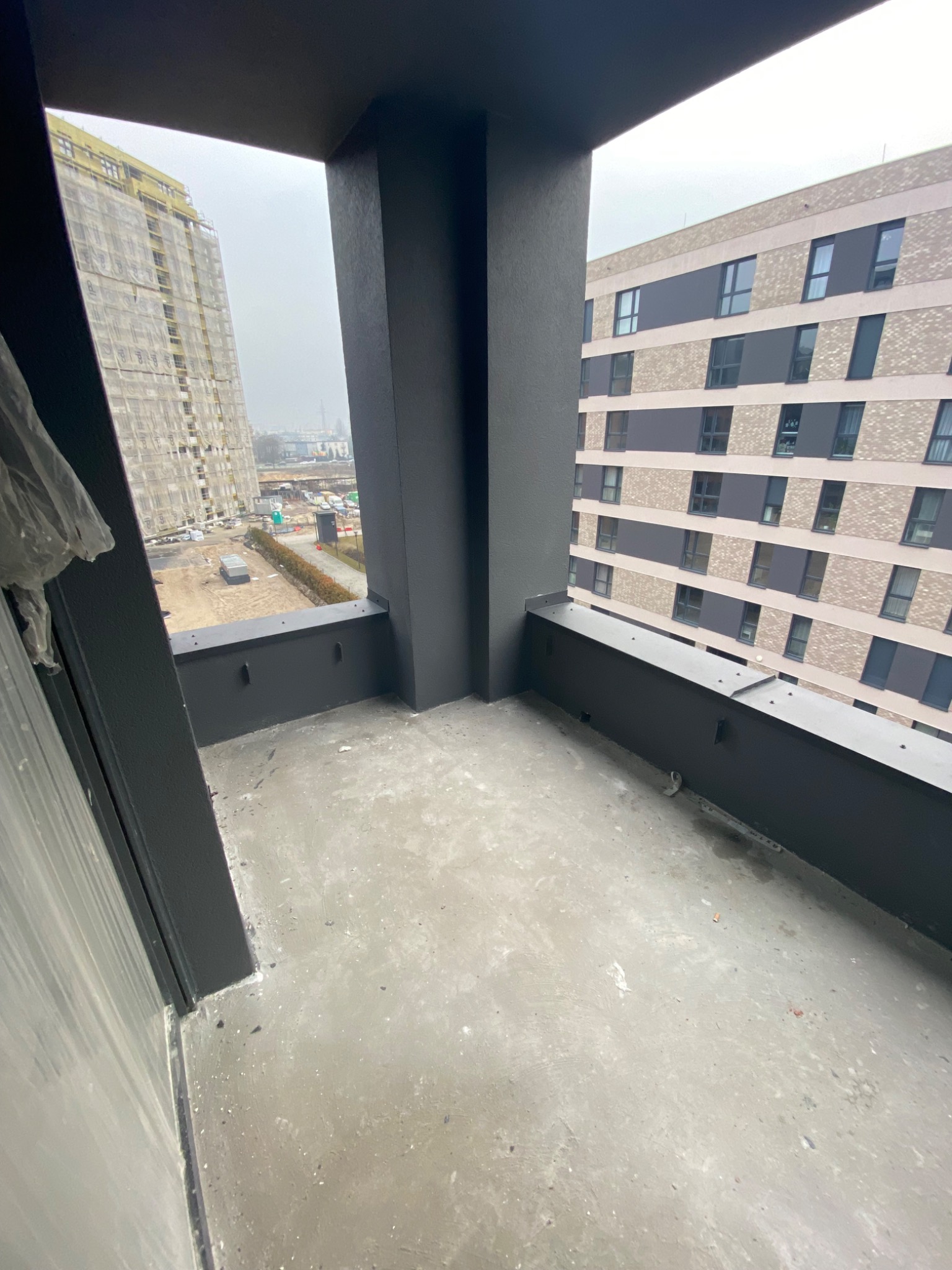 Surowy betonowy balkon w nowym budownictwie z widokiem na plac budowy i sąsiedni budynek mieszkalny w pochmurny dzień.