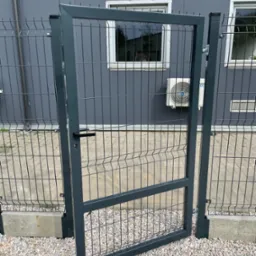 Otwarta, szara furtka z metalowej siatki panelowej, widoczna na tle szarego budynku z oknami i klimatyzatorem, osadzona na betonowym fundamencie i żwirowym podłożu.