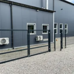 Nowoczesne ogrodzenie z metalowych paneli na tle szarego budynku przemysłowego z klimatyzatorami i oknami.