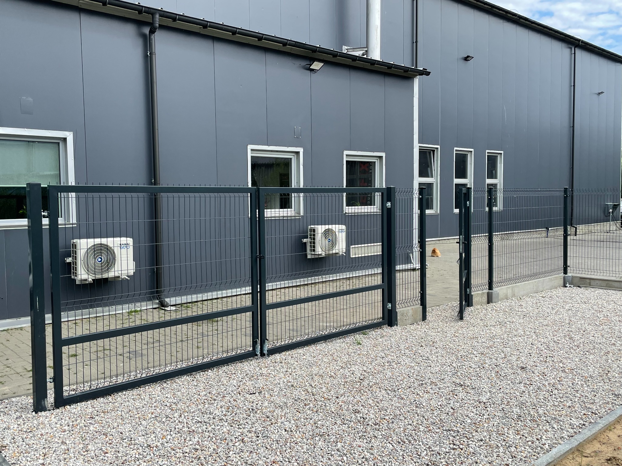 Nowoczesne ogrodzenie z metalowych paneli na tle szarego budynku przemysłowego z klimatyzatorami i oknami.