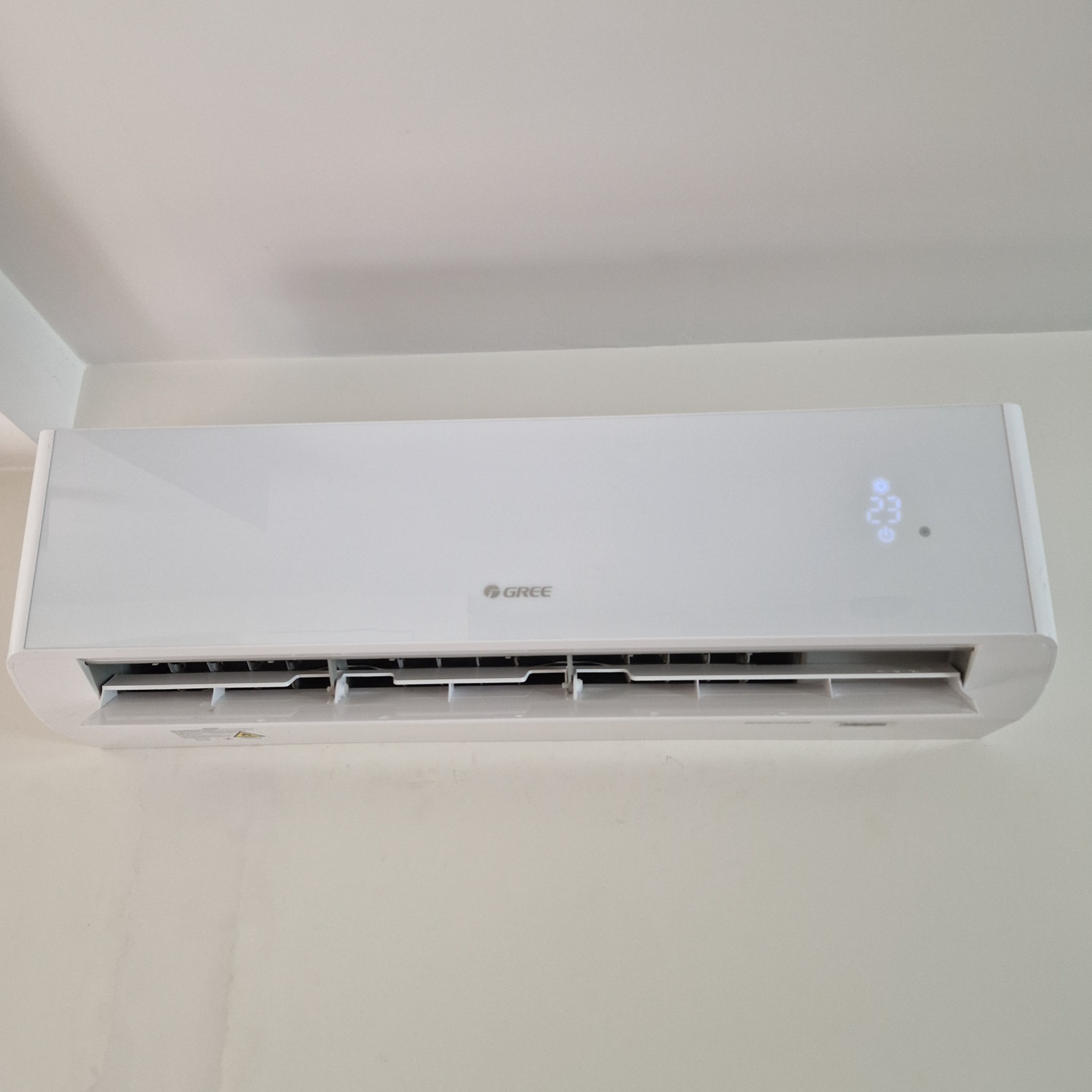 Biała jednostka klimatyzacji marki Gree zamontowana na ścianie, wyświetlająca temperaturę 23 stopnie Celsjusza.