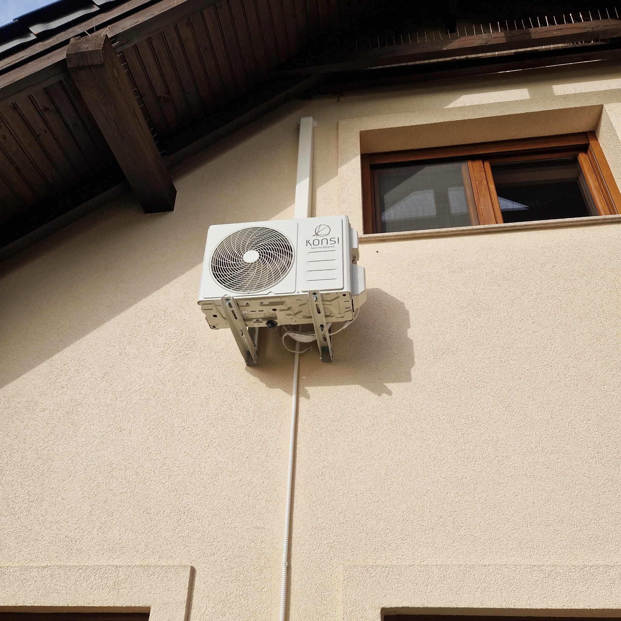 Biała jednostka zewnętrzna klimatyzacji marki Konsi zamontowana na beżowej elewacji domu, widoczne wsporniki, rura odprowadzająca oraz fragment okna z brązową ramą i zabezpieczenia przeciw ptakom...