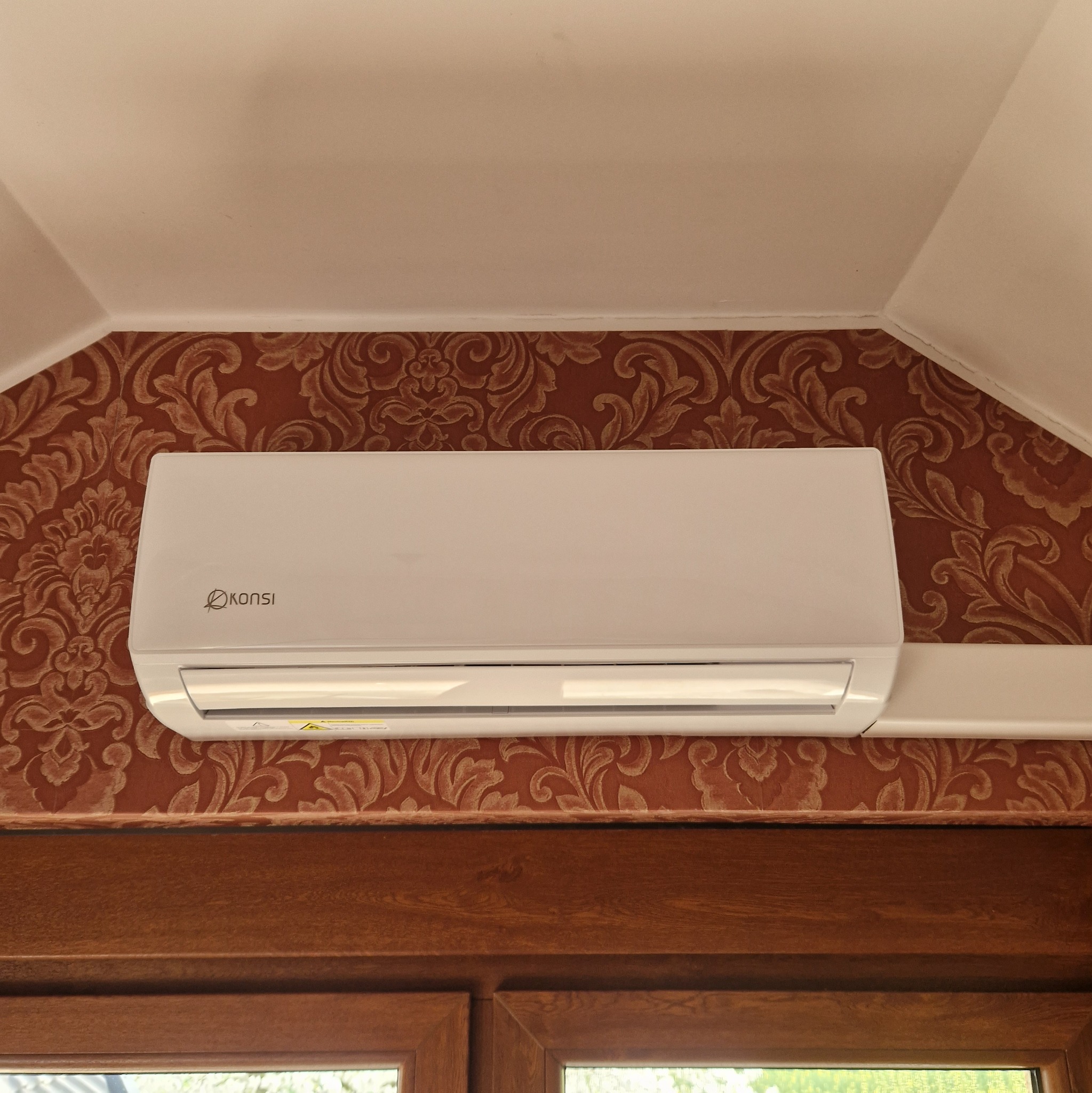 Biały klimatyzator marki Konsi zamontowany na ścianie z wzorzystą tapetą w pomieszczeniu z drewnianą ramą okienną u dołu kadru.
