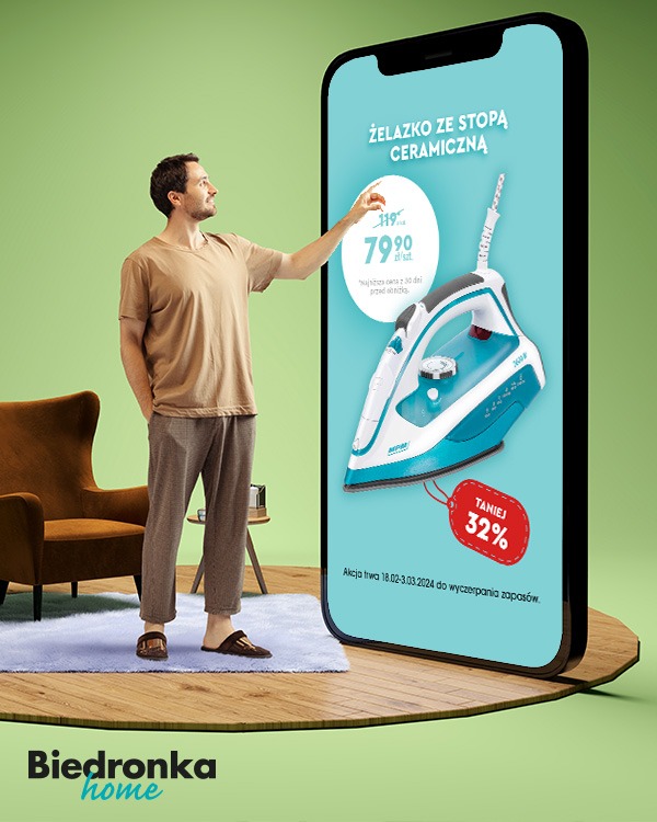 Mężczyzna w domowym stroju dotyka palcem ekranu smartfona, na którym wyświetlona jest reklama żelazka z ceramiczną stopą w promocyjnej cenie, obok brązowy fotel i dywanik na drewnianej podłodze.