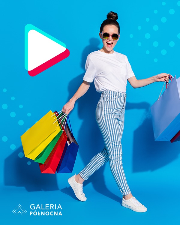 Uśmiechnięta kobieta w okularach, w ruchu, niosąca kolorowe torby na zakupy na tle błękitnego tła z logo galerii.