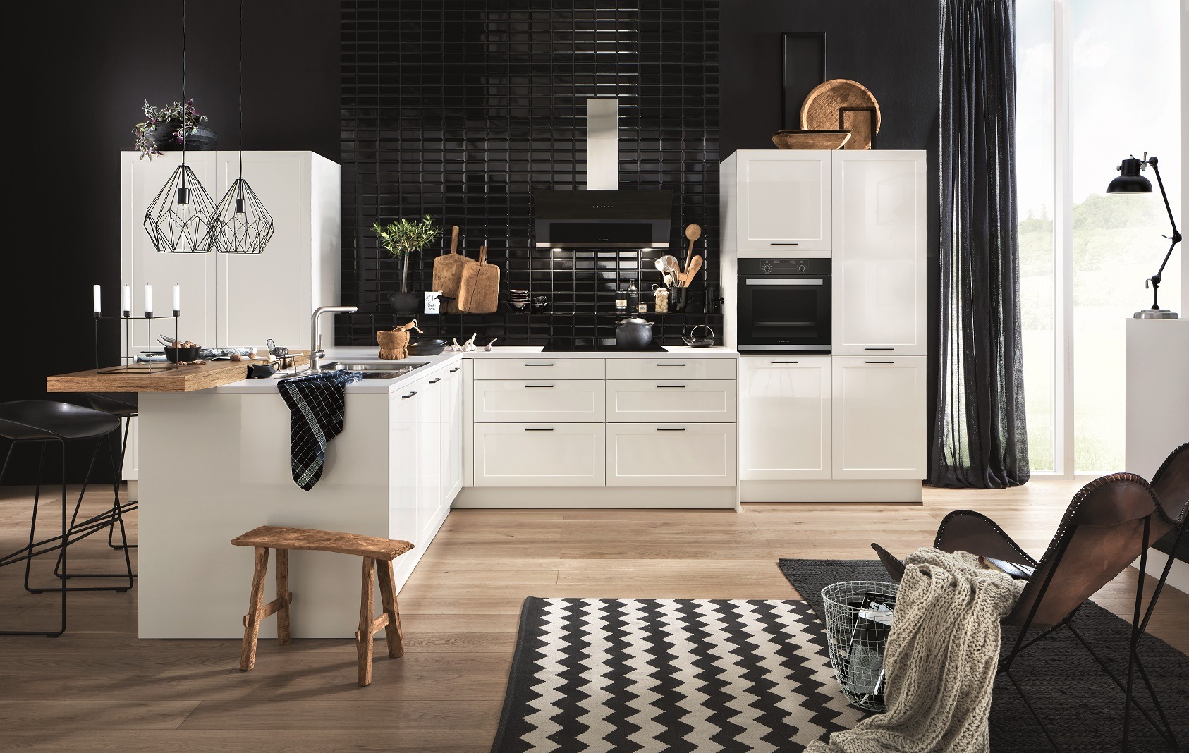 Biała, nowoczesna kuchnia z czarnymi akcentami: płytki, okap, dekoracje. Drewniane blaty, minimalistyczne lampy i wzorzysty dywan dodają charakteru.