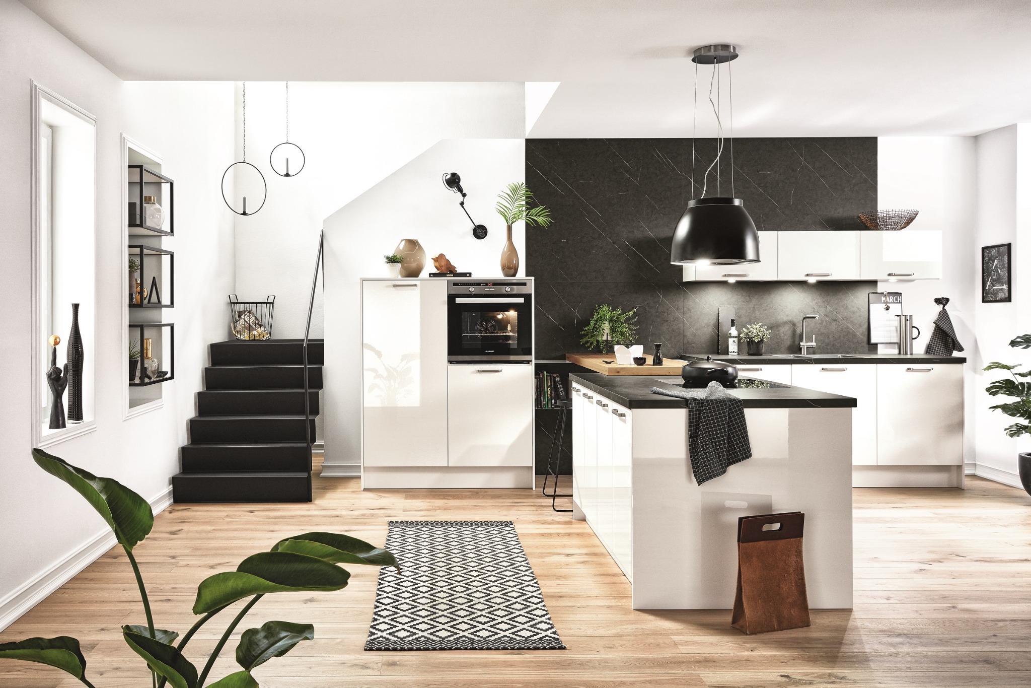 Nowoczesna, biała kuchnia z czarnym blatem i schodami. Lampa wisząca, rośliny doniczkowe i dywanik dodają wnętrzu charakteru. Minimalistyczny design z akcentami.