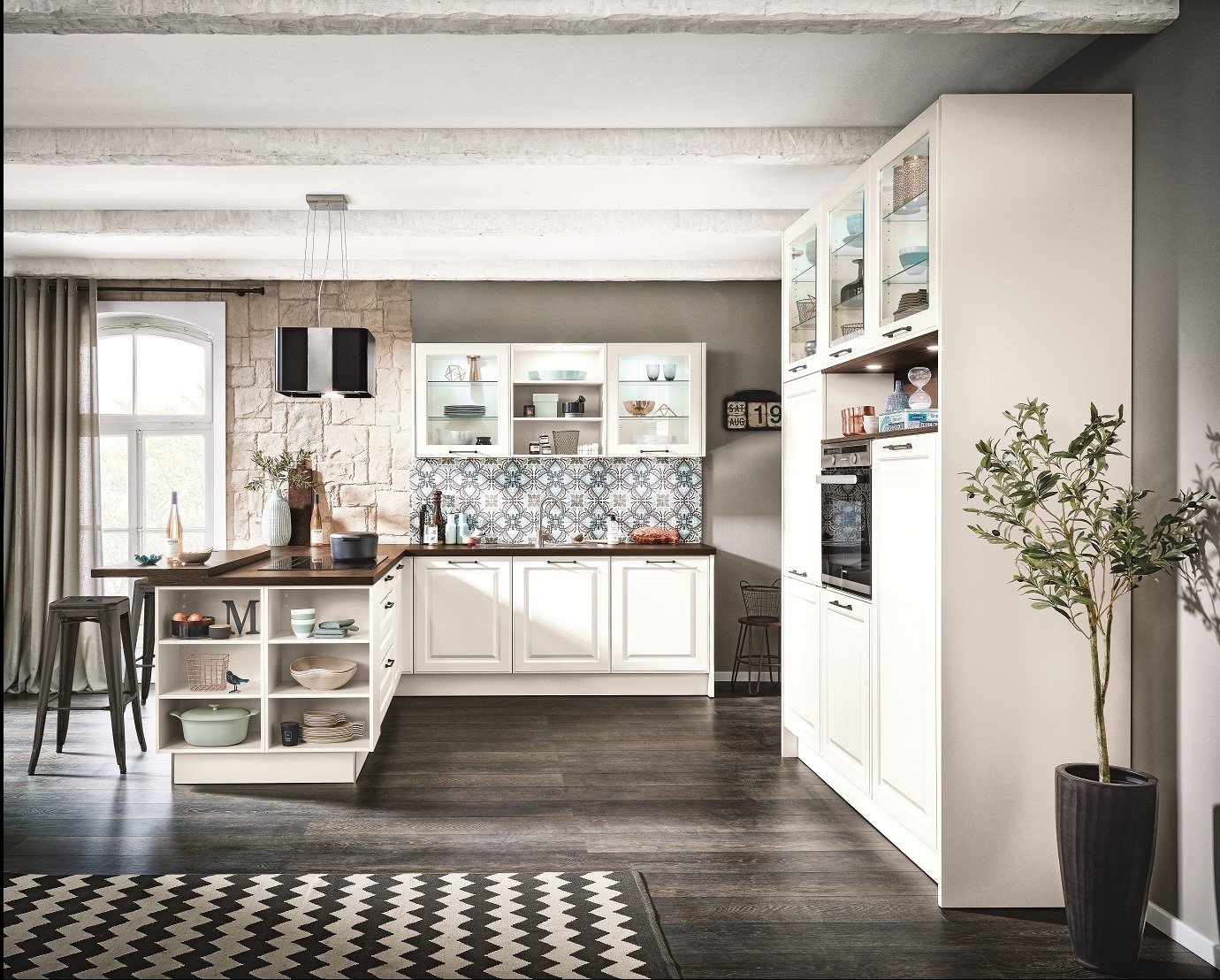 Jasna kuchnia w stylu rustykalnym z wyspą i wzorzystą mozaiką nad blatem. Widoczne przeszklone szafki i czarny okap. Na pierwszym planie roślina w doniczce.